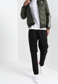 Olivově zelená prošívaná bomber bunda s bílými pruhy, kombinovaná s černými kalhotami a bílými teniskami. Má kulatý výstřih a zapínání na zip.