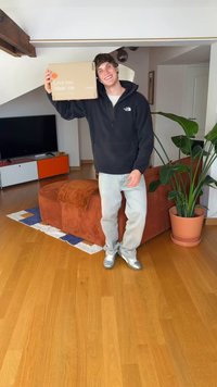 Caixa de cartão segurada por uma pessoa vestindo um casaco com capuz preto e jeans claros. Ao fundo, há um sofá de veludo cotelê castanho e uma planta verde.