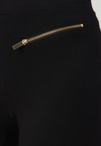 Leggings noirs avec une texture lisse, dotés d'une poche zippée dorée sur le côté gauche. La coupe est ajustée et élégante.