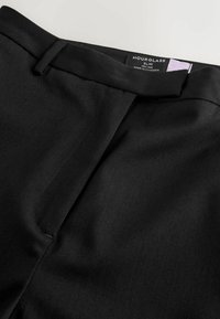 Pantalon noir slim taille haute avec une texture lisse, des coutures visibles et une étiquette de ceinture sur mesure indiquant "Hourglass".