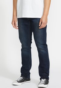 Unos vaqueros de mezclilla azul oscuro con un corte ajustado, una textura sutil, combinados con unas zapatillas negras con cordones blancos y suelas de goma.