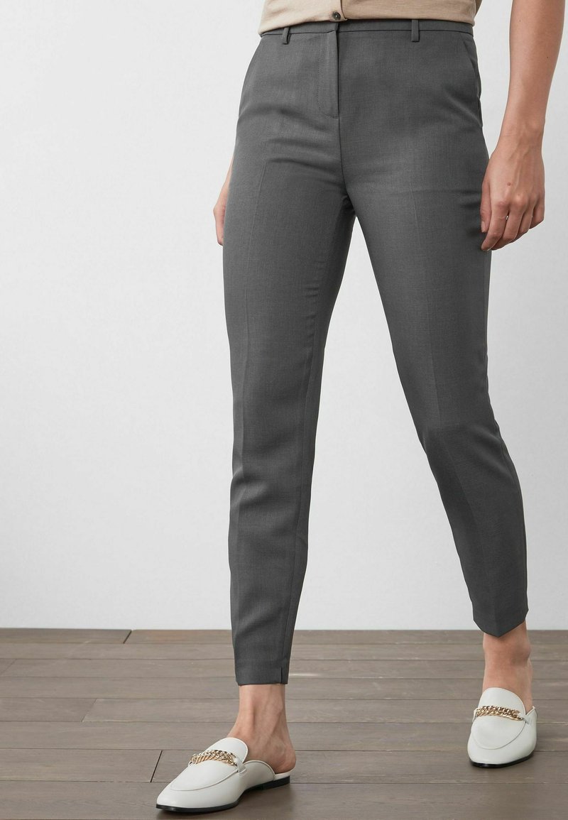 Personne portant un pantalon gris foncé ajusté et des mocassins blancs sans lacets avec un détail chaîne dorée, debout sur un sol en bois devant un fond uni.