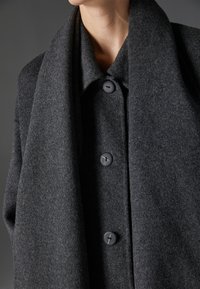 Abrigo de lana de color gris oscuro con un gran collar y cinco botones en la parte delantera. Textura suave con una silueta estructurada y diseño minimalista.