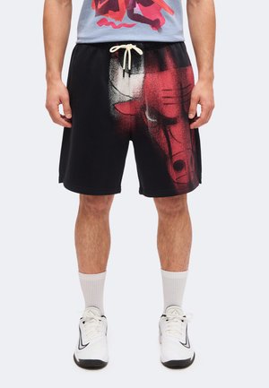 Homme portant un short noir avec un logo rouge des Chicago Bulls, un cordon blanc, des chaussettes blanches et des chaussures de sport blanches, debout devant un fond uni.