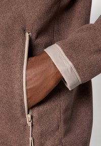 Veste en laine marron avec une poche zippée, dotée d'un poignet en tissu clair contrastant. La texture est douce, et le design est simple et fonctionnel.