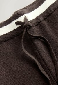 Bruine gebreide joggingbroek met een tailleband met witte strepen, voorzien van een trekkoord dat met een knoop is vastgemaakt. Zachte textuur en elastische afwerking.