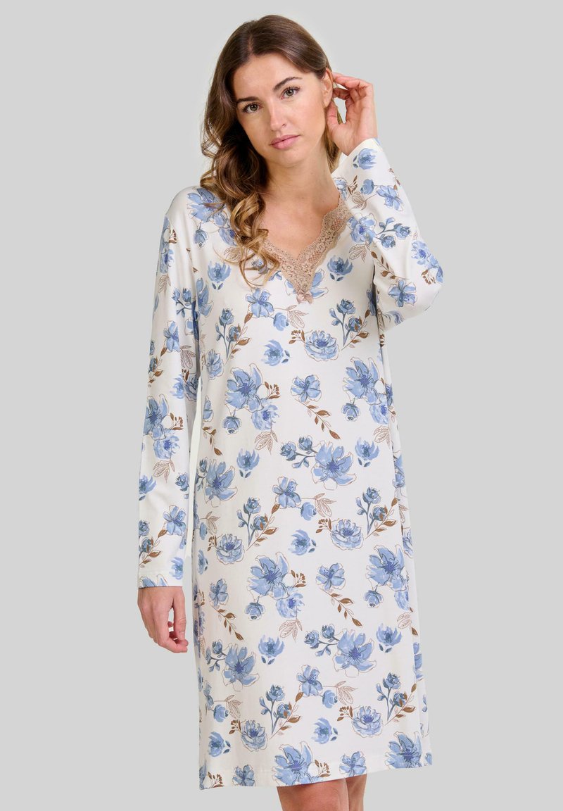 Mujer con cabello ondulado que lleva una camisa de dormir blanca de manga larga con estampados florales azules y ribete de encaje, de pie contra un fondo simple.