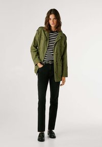 Olīvu zaļš militārā stila jaka ar vairākiem kabatām, melnas un baltas svītrainas blūze, melnas šauras džinsas, un melnas klājējzābaki.