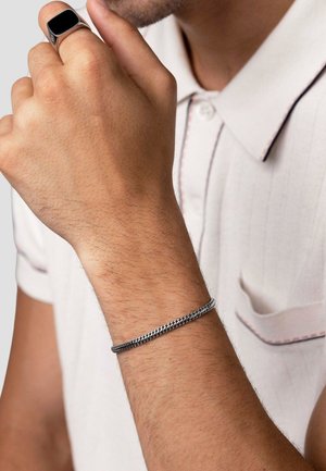 Silver chain bracelet on wrist, featuring a woven design. The wearer is also shown wearing a black ring and a light-colored collared shirt.