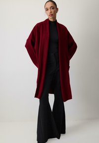 Nő, aki egy hosszú bordó kardigánt visel fekete magas nyakú felső és bő szárú fekete nadrág felett, egyszerű világos háttér előtt áll.