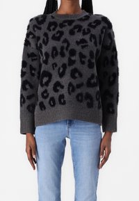 Maglione grigio a maglia con macchie leopardate nere, caratterizzato da un colletto e polsini a costine. Tessuto morbido con una vestibilità comoda. Indossato con jeans blu.