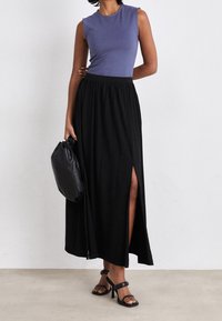 Žena v tílku fialové barvy bez rukávů a černé sukně s vysokým pasem a rozparkem na stehně, držící černou kabelku, v černých sandálech na vysokém podpatku.