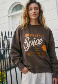 Noor noor, kellel on lainelised blondid juuksed, kannab pruuni sviitrit, millel on tekst "Pumpkin Spice Martini Cocktail" ja kõrvitsate illustratsioonid.