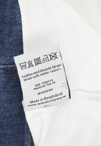 Biała metka przymocowana do dżinsu, zawierająca instrukcje prania, nazwę marki "Seaside Company" oraz szczegóły "Wyprodukowane w Bangladeszu".