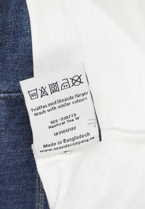 Pflegeetikett auf weißem Stoff mit Denim, zeigt Waschsymbolen und Text, einschließlich "Mit ähnlichen Farben waschen" und "Hergestellt in Bangladesch."