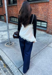 Žena s dlhými tmavými vlasmi kráčajúca po mestskom chodníku, oblečená v čiernom topu pod bielou krajkovou kamarátkou a širokých tmavomodrých džínsoch, drží hnedú tašku.