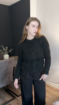 Zwarte, gestructureerde cardigan met knopen en een trekkoord. Draagt over zwarte broek met wijde pijpen. Neutrale achtergrond met planten in een pot.