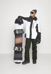 Mees musta ja valge snowboardi jope, mustade pükste, kinnaste ja mütsiga seisab, hoides snowboardi püsti ühtlase tausta ees.