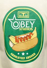 Λογότυπο Obey Studios με ψωμάκι για χοτ ντογκ που κρατά μια μπανάνα καλυμμένη με κέτσαπ, πλαισιωμένο σε πράσινο και κίτρινο, με λεζάντα «Απόλυτα παράλογο.»