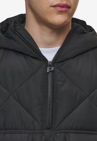 Giacca nera trapuntata con zip frontale, cappuccio e finitura strutturata. Presenta un motivo di cucitura diagonale e robusti accessori per la zip.
