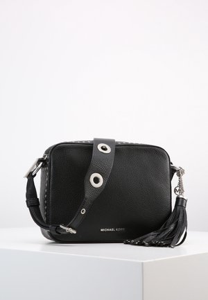 Czarna skórzana torebka crossbody z szerokim paskiem, z dużymi srebrnymi oczkami, ozdobą z chwostem oraz srebrnym logo Michael Kors z przodu.