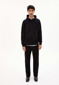 Haut à capuche en coton noir avec cordon, poche avant, associé à un pantalon noir et des bottes en cuir. Superposé sur une chemise à carreaux bleu clair.