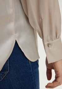 Beige, doorschijnende blouse met lange, volumevolle mouwen en een afgeronde zoom. Heeft een decoratieve knoop op de manchet. Draagt over donkere, taps toelopende jeans.