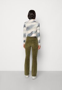 Vrouw met kort donker haar staat met haar rug naar de camera, gekleed in een lange mouwen shirt met wolkenpatroon, olijfgroene corduroy broek en witte sneakers.