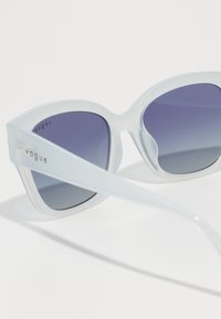 Ochelari de soare tip cat-eye cu o ramă albă transparentă și lentile violet cu degradé. Detaliu gravat "vogue" pe braț. Textură netedă.