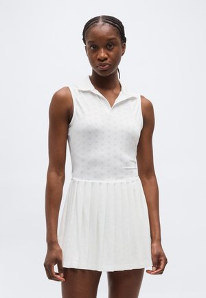 Žena s vrkočmi, oblečená v bielych bezrukávových tenisových šatách s plisovanou sukňou na jednoduchom svetlom pozadí.