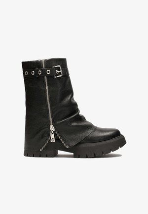 Botte mi-mollet en cuir noir avec semelle à relief épais, fermeture éclair latérale partiellement ouverte, et sangle décorative à boucle près du haut.