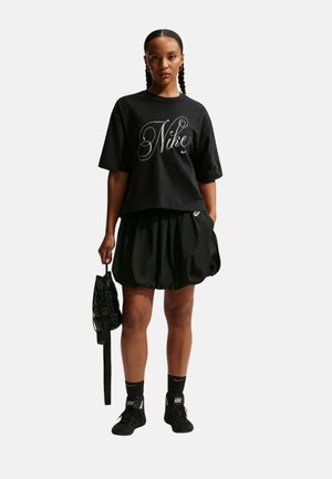 Mujer con el cabello trenzado que lleva una camiseta negra de Nike, falda negra, calcetines negros de Nike y zapatos, sosteniendo una bolsa negra con textura a su lado.