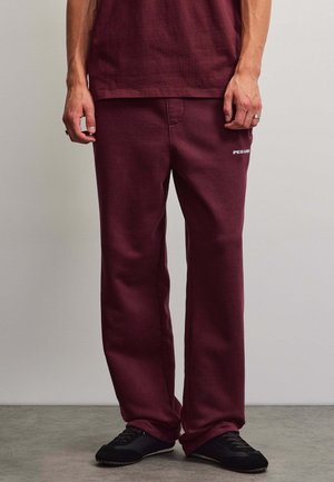 Man wearing burgundy sweatpants and matching shirt with black sneakers standing on gray floor against light wall.
