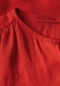 Tessuto di cotone rosso con una consistenza morbida, caratterizzato da un collo a barchetta e leggeri plissettami nella cucitura della spalla.