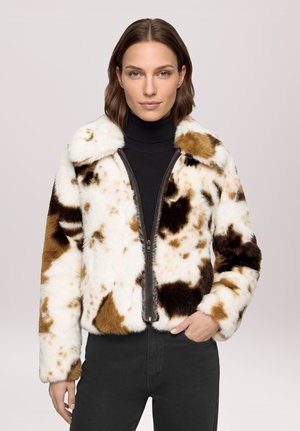 Femme portant une veste en fausse fourrure marron et blanche à taches, un col roulé noir, un pantalon noir et des bottines noires à talons, debout avec une main dans la poche.
