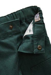 Tissu en chevrons vert foncé avec une taille élastique. Comprend des accessoires en métal brun et une étiquette blanche à l'intérieur.