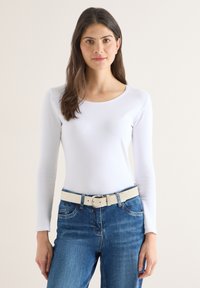 Jeune femme aux longs cheveux foncés portant une chemise blanche à manches longues et un jean bleu avec une ceinture beige, se tenant devant un fond uni.