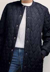 Giacca trapuntata blu navy con collo rotondo, chiusura con bottoni a pressione e tasche laterali, abbinata a una camicia bianca e jeans blu.