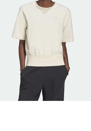 Personne portant un sweat-shirt beige à manches courtes et col ras du cou avec un ourlet côtelé, un pantalon gris foncé, les mains dans les poches, sur un fond uni.