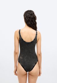 Γυναίκα με μακριά σγουρά μαλλιά, φορώντας μια μαύρη, ριγωτή ολόσωμη φόρμα χωρίς μανίκια, εμφανιζόμενη από πίσω σε απλό λευκό φόντο.