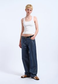 Vit ruggad mastectop kombinerad med mörkblå vida denimjeans, accentuerad med en svart hjärtformad bältesspänne och bruna mönstrade skor.
