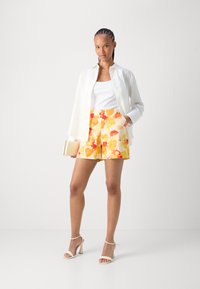 Camicia bianca a bottoni, canotta bianca a coste, pantaloncini floreali arancioni e gialli, tacchi abbinati, tiene in mano una clutch dorata.