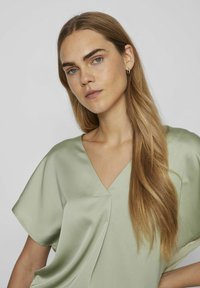 Top in satin verde chiaro con scollatura a V e maniche corte e ampie, caratterizzato da una texture liscia e un delicato drappeggio.