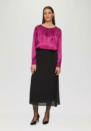 Frau in einer leuchtend lila seidenen Bluse, schwarzem plissiertem Midirock, schwarzen transparenten Strumpfhosen und schwarzen hohen Schuhen, die vor einem schlichten Hintergrund steht.