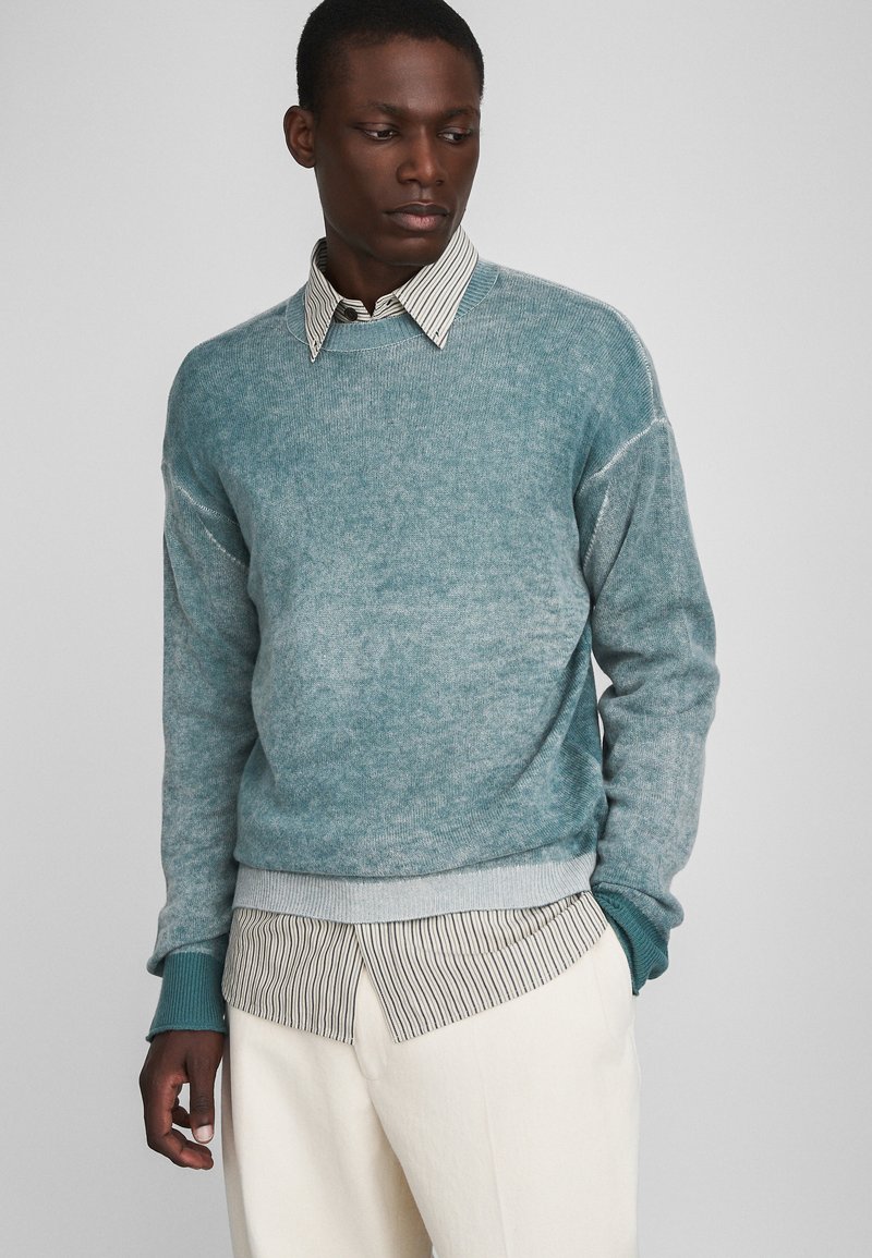 Heleheles sinine ümar kaelusega sviiter, mis on valmistatud pehmest kudumist, on kihiliselt tükeldatud triibulise kraega särgi peale, millega on kokku sobitatud kreemika värviga püksid.