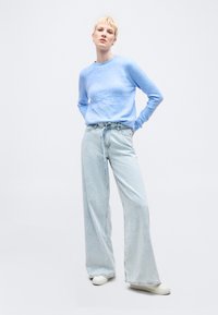 Γυναίκα με κοντά ξανθά μαλλιά που φοράει ανοιχτό μπλε πουλόβερ, τζιν με ψηλή μέση και φαρδιά μπατζάκια με ζώνη, και λευκά αθλητικά παπούτσια σε λευκό φόντο.