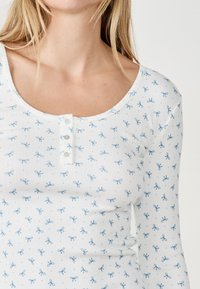 Witte lange mouwen top met blauwe strikprint, voorzien van een ronde halslijn en een detail met drie knopen aan de voorkant. Soepele textuur.