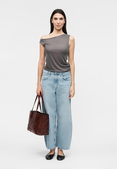 Γυναίκα με μακριά σκούρα μαλλιά που φορά ασύμμετρο γκρι τοπ, φαρδιά ανοιχτό μπλε τζιν, μαύρες μπαλαρίνες, κρατώντας πλεκτή καφέ δερμάτινη τσάντα tote.