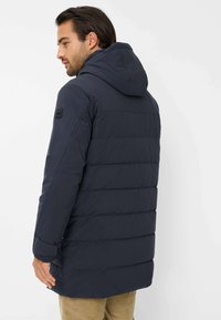 Giacca imbottita blu scuro con cappuccio, caratterizzata da un design strutturato e trapuntato, polsini elasticizzati e una patch sulla spalla sinistra.