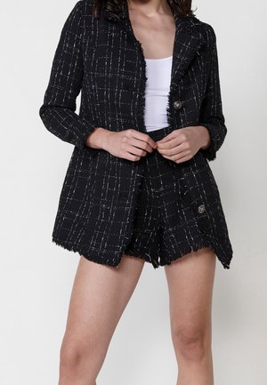 Kobieta ubrana w czarno-białą, kratkowaną teksturowaną marynarkę i dopasowane szorty, na białym topie, trzymająca ręce na przodzie marynarki.